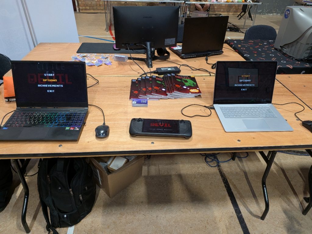 Little Devil setup at the Rapture Gaming Festival in Colchester, running on 2 laptops and a Steam Decki, with comics and business cards on the table